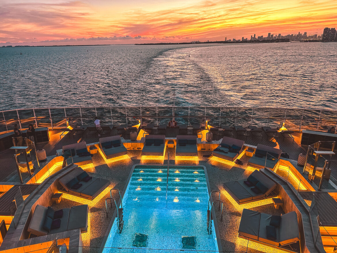 A luxurious cruise ship deck at sunset, featuring a central swimming pool surrounded by illuminated lounge chairs. The ocean stretches out to the horizon, with a city skyline visible in the distance under a vibrant, colorful sky.