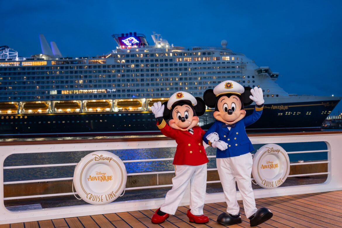 Two costumed characters, resembling iconic cartoon mice, are dressed as ship captains in red and blue uniforms. They are standing on a deck with a large cruise ship in the background. The ship has the words "Disney Adventure" and "Disney Cruise Line" visible on life preservers attached to the railing. The scene is set during twilight, with the ship's lights illuminating the area.