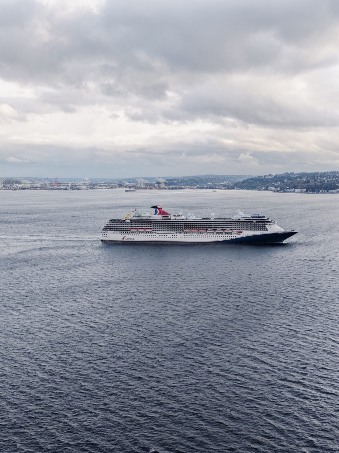 A large cruise ship is sailing on a calm body of water under a cloudy sky. In the background, there is a distant shoreline with industrial structures and hills.