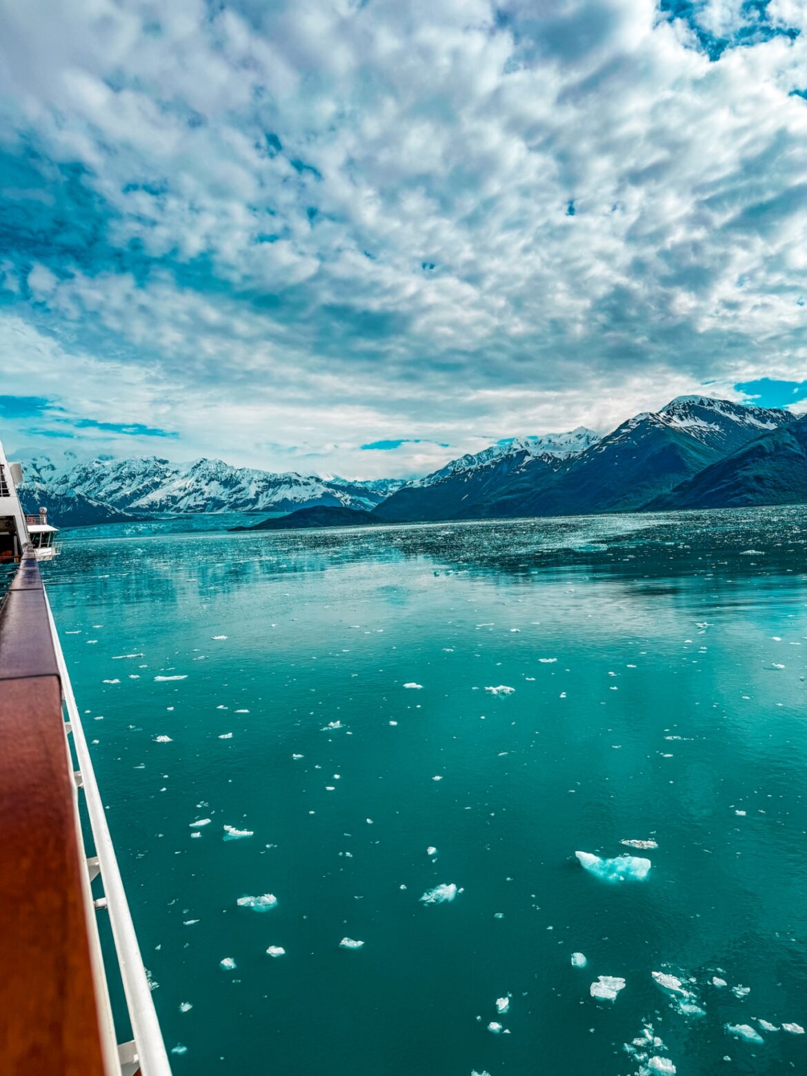 A scenic view from a ship's deck shows a vast expanse of turquoise water with small ice chunks floating on the surface. In the background, there are snow-capped mountains under a partly cloudy sky. The ship's railing is visible on the left side of the image.